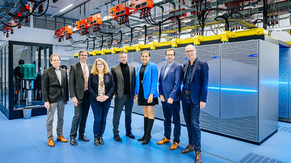 Five men and two women, all smartly dressed, stand in a computer centre and smile.