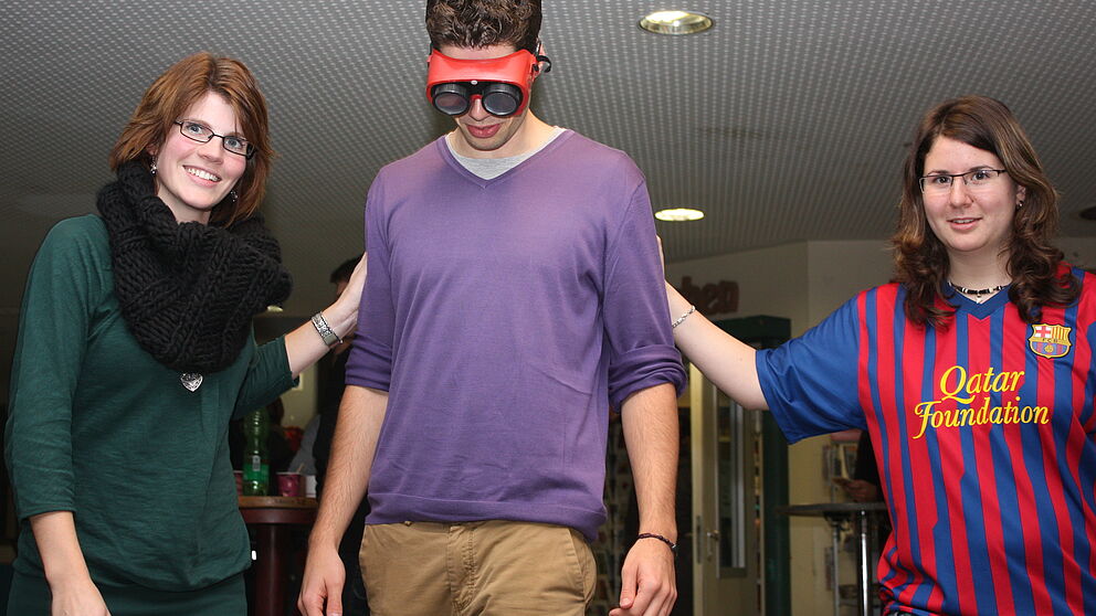 Foto (Universität Paderborn, Patrick Kleibold): Mit den Augen eines Angetrunkenen: Henrik Jahns versuchte mit der Suchtbrille einer Linie zu folgen. Nele Tiedemann (links) und Isabell Schwartz mussten ihm dabei behilflich sein.