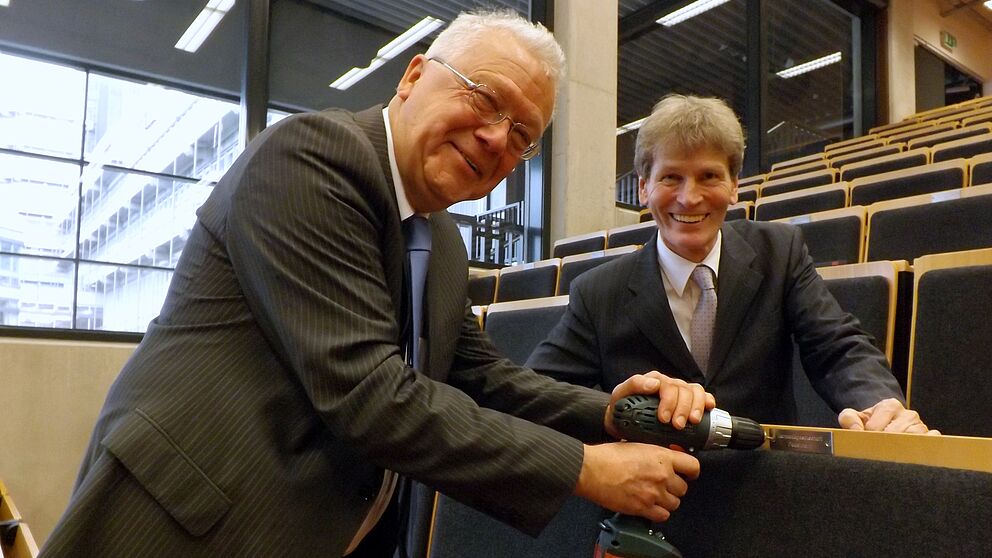 Foto (Universität Paderborn, Jasmin Rostam): Dr. Rolf Dietz von der Harmoniegesellschaft (li.) bringt die Plakette am gestifteten „Leerstuhl“ an. Prof. Dr. Nikolaus Risch freut sich über die Spende.