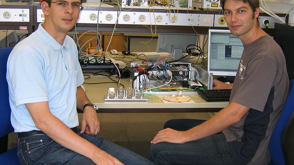 Foto (Universität Paderborn): V. li.: Dr.-Ing. Vitali Mirvoda und Dipl.-Ing. Benjamin Koch arbeiten an ihrem Versuchsaufbau zur unterbrechungsfreien Polarisationsregelung.