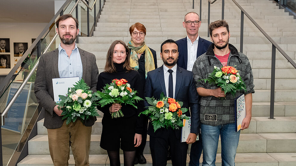 Zwei Frauen und vier Männer stehen in zwei Reihen auf einer Truppe, vier von Ihnen halten Blumensträuße.