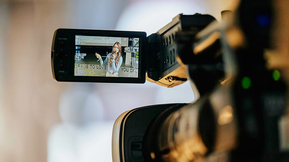In the foreground a camera, on whose screen you can see a woman explaining something.