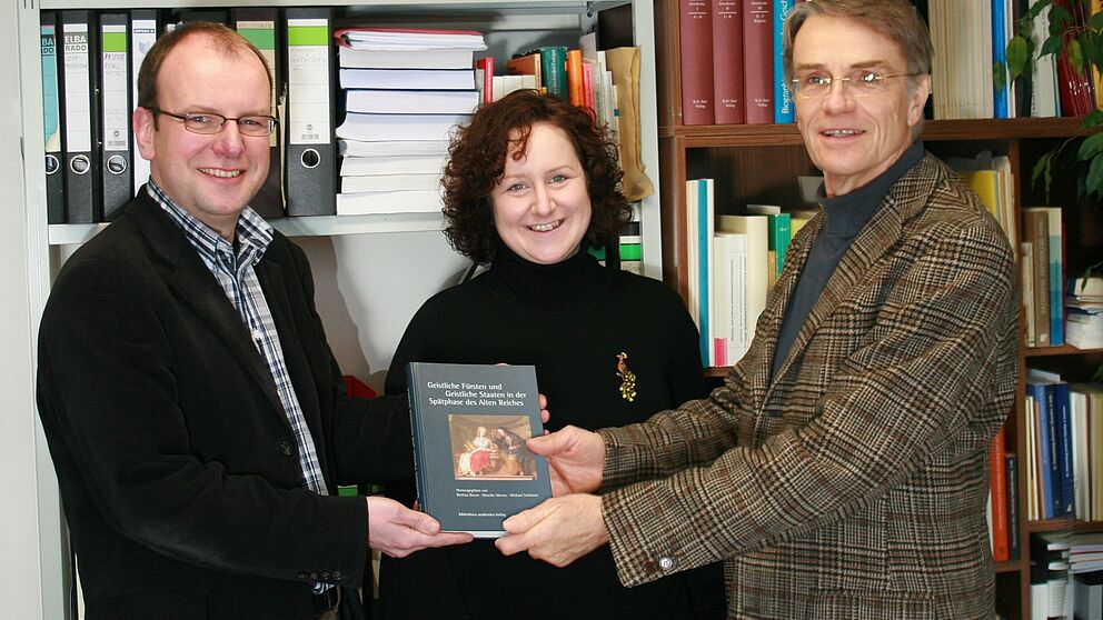 Foto (Dirk Leyhe, Universität Paderborn): V. li.: Herausgeber des Tagungsbandes Dr. Michael Ströhmer und Dr. Mareike Menne überreichen Prof. Dr. Frank Göttmann die jüngste Bilanz zum Paderborner Forschungsunternehmen „Geistliche Staatlichkeit im Al
