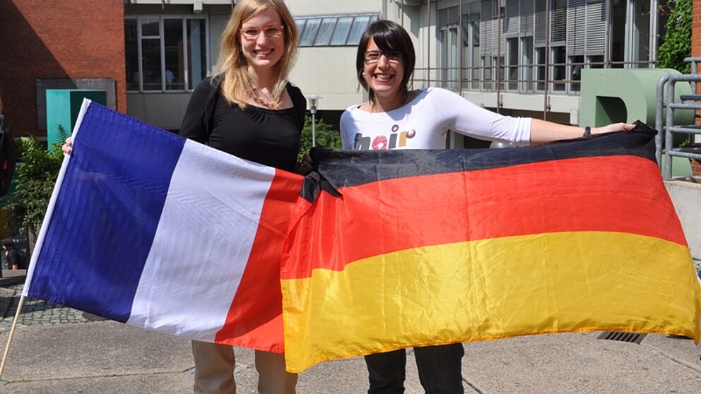 Foto (Universität Paderborn, Mark Heinemann): Europäische Studien: Symbolisch halten die Kommilitoninnen Alexandra Weckwerth (li.) und Adeline Besnier die Landesflagge des jeweils anderen Partnerlandes.