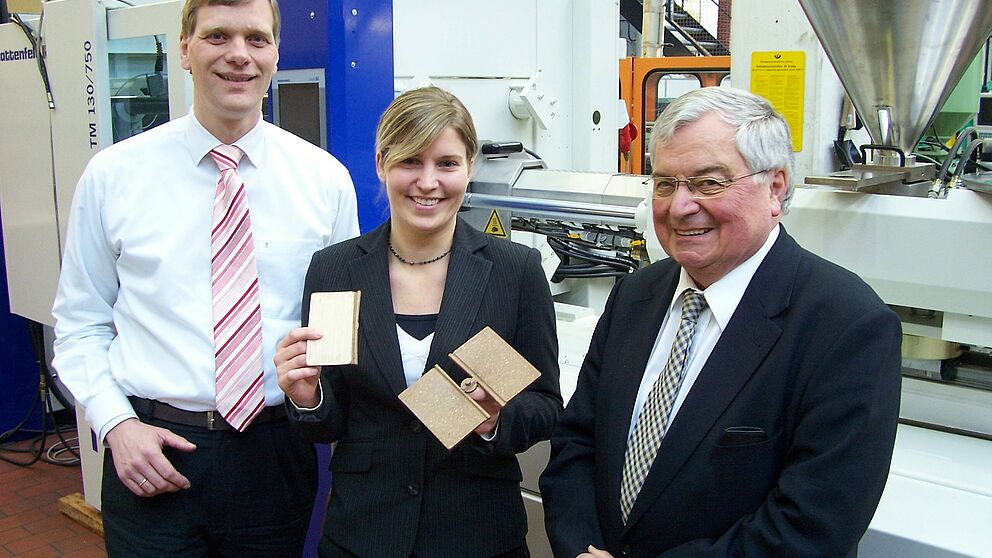 Foto (Mark Heinemann): Holz und Kunststoff (v. l.): Prof. Dr.-Ing. Volker Schöppner, Cathrin Funke, hier mit gefertigten Modellen aus dem Forschungsprojekt, und Institutsleiter Prof. Dr.-Ing. Helmut Potente vor einer Spritzgussmaschine, die aktuell im Ve