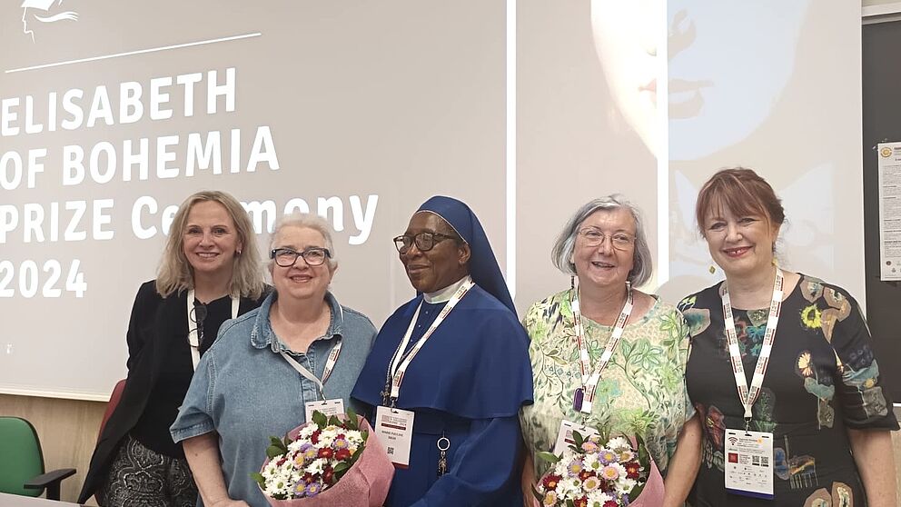 Fünf Frauen stehen nebeneinander und lächeln, zwei von ihnen halten jeweils einen Blumenstrauß.