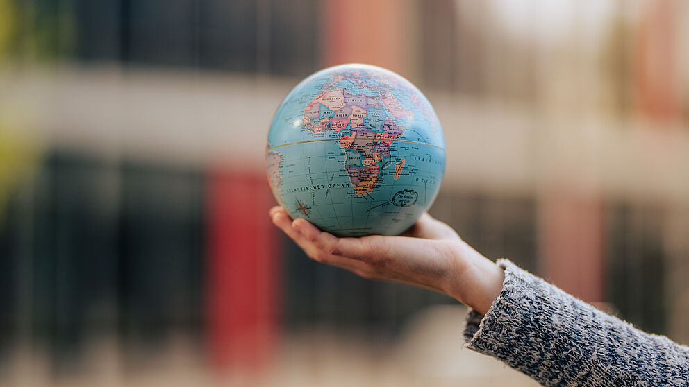 Ein kleiner Globus wird von einer Hand gehalten, im Hintergrund unscharf das Gebäude I (Symbolbild für Internationales Profil)