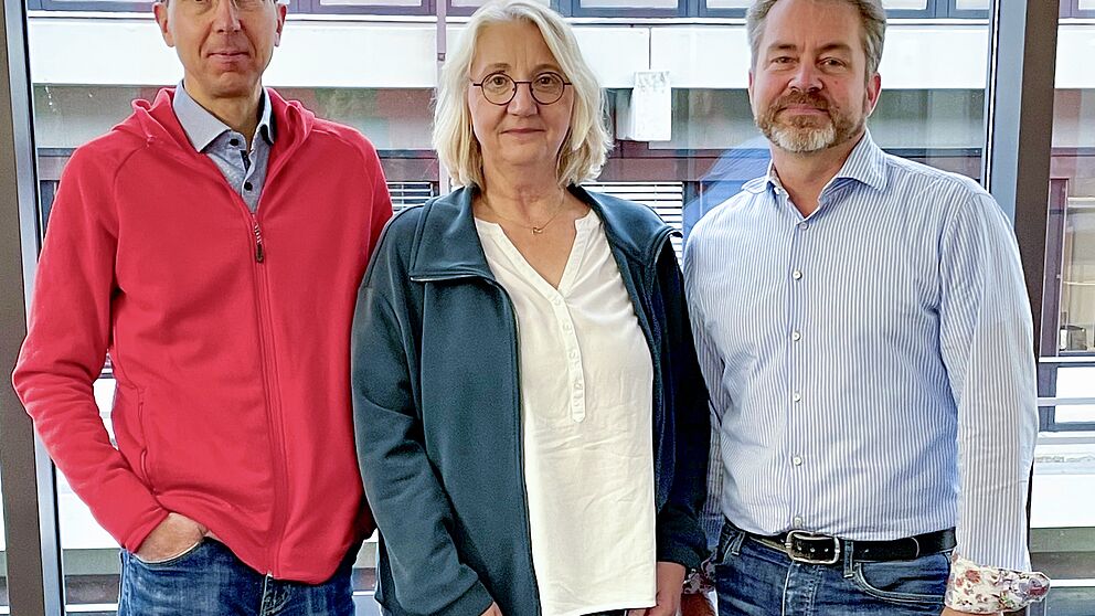 Eine Frau und zwei Männer stehen drinnen vor einer Fensterfront.