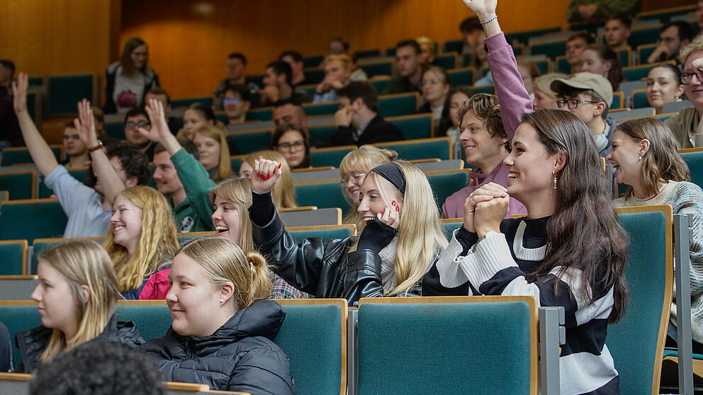 Eine Gruppe von jungen Menschen sitzt im Hörsaal, eine heben die Hand und die Arme und sie lächeln. 