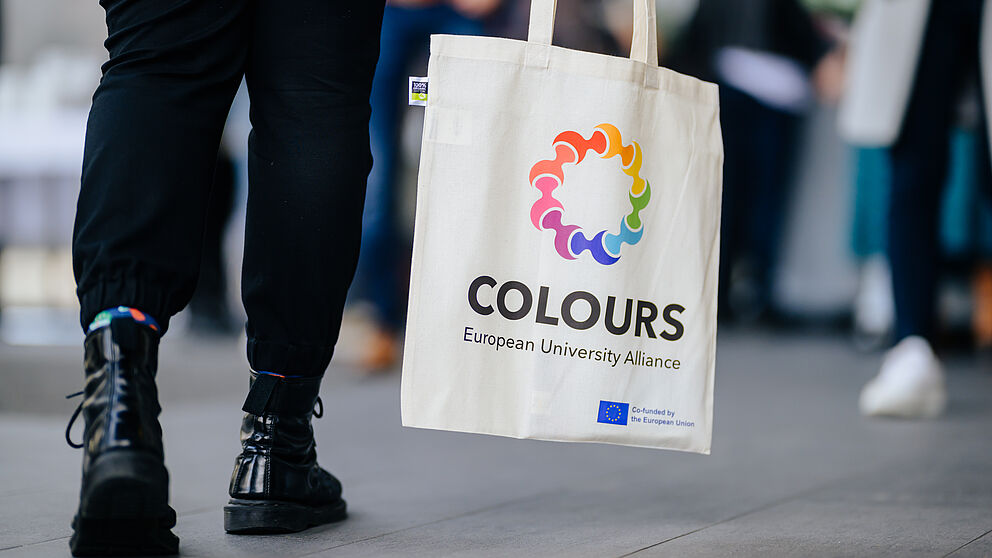 Legs in black trousers and black boots, next to a bag with the logo of the European university alliance COLOURS printed on it. 