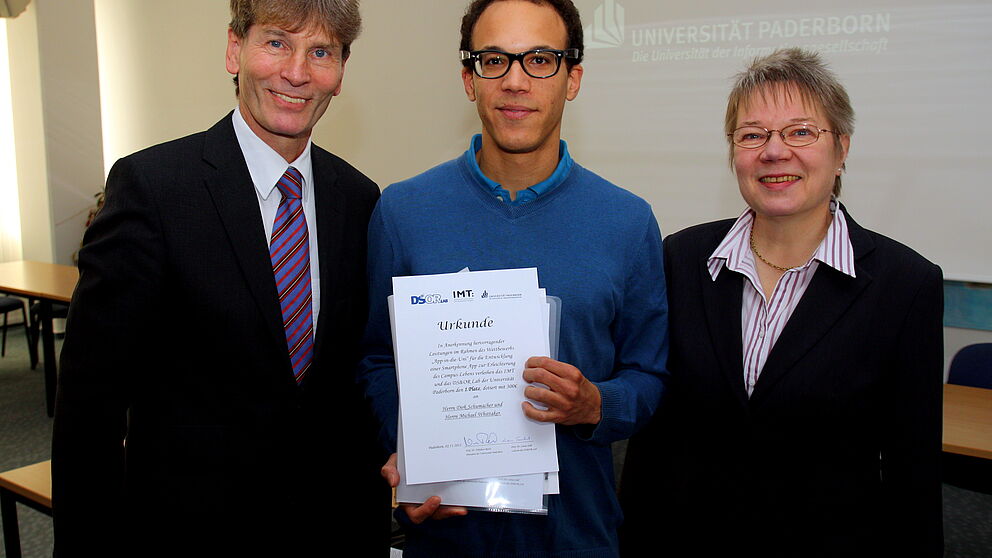 Foto (Universität Paderborn, Patrick Kleibold): Prof. Dr. Nikolaus Risch und Prof. Dr. Leena Suhl überreichen die Urkunde für den ersten Preis im Wettbewerb „App in die Uni“ an Michael Whittaker für dessen App „I-UPB“.