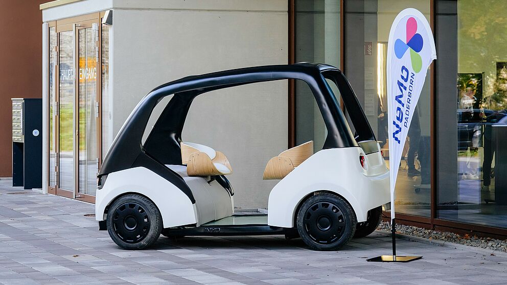 In the foreground, a so-called "Cab", a model vehicle with which people are to be sustainably mobile in the "New Mobility Paderborn" project. Next to it is a flag with the project logo.